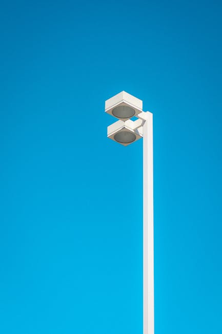 A tall white street lighting pole with two large, rectangular, modern light fixtures mounted near the top, positioned outdoors against a clear blue sky. The pole is situated on a paved surface, likely a driveway or sidewalk, and is part of urban street infrastructure. Although no furniture, boxes, or moving equipment are visible, the setting suggests a location where loading or unloading may occur during house removals. The bright sunlight, absence of shadows or additional objects, and the clean, minimalistic design of the lighting fixtures reflect a contemporary environment. This image could be used to illustrate the area around a home or property involved in a furniture transport or moving process, aligning with the theme of home relocation services provided by companies like Removal Van Kingston, especially related to avoiding hidden removal charges in Kingston.