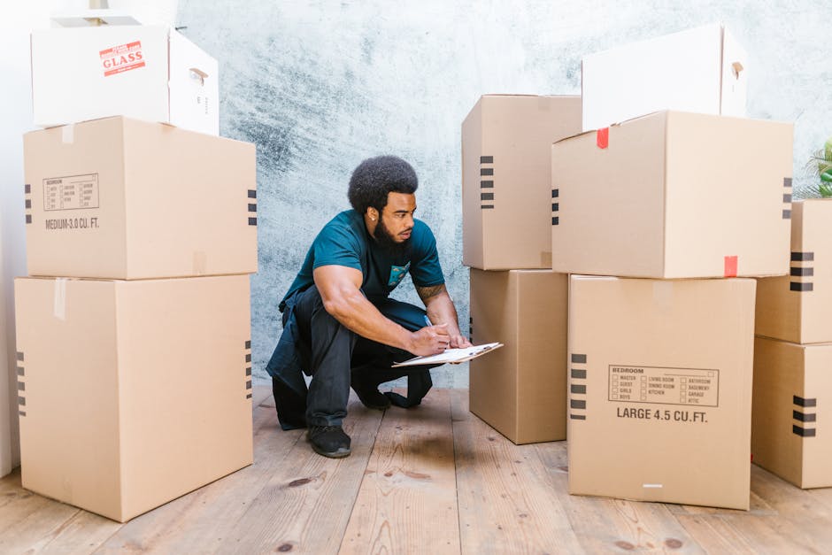 A man in casual clothing is kneeling on a wooden floor indoors, surrounded by large cardboard moving boxes of various sizes, some with printed labels indicating contents such as bedroom furniture and large capacity. He is holding a clipboard and appears to be inspecting or marking the boxes as part of the home relocation process. The boxes are packed with packing materials like paper and plastic wrap, and some are secured with packing tape or red straps. Behind him, a textured wall and a closed door are visible, with additional smaller boxes stacked nearby. This scene reflects the packing and loading stages of a house move, supported by a professional removal service such as Removal Van Kingston, which ensures organized furniture transport and efficient packing during residential relocations.