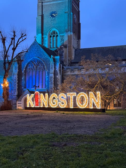 A large illuminated sign spelling 'KINGSTON' in capital letters is positioned on the ground in front of a historic church building at dusk. The sign features multicoloured lights, with the letter 'I' in red and others in white. The church, constructed from stone with Gothic-style pointed windows, is lit in blue, highlighting its architectural details. To the left of the sign, there is a leafless tree, and to the right, a tree with some foliage. The area around the sign appears to be a paved or gravel surface, with patches of grass in the foreground. This scene is related to house removals and logistics support by Removal Van Kingston, illustrating a typical exterior setting during a home relocation or furniture transport process in Kingston.