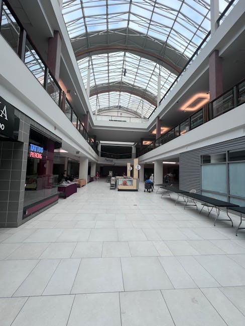 An interior view of a spacious shopping mall atrium with a high, curved glass roof allowing natural light to illuminate the area. The ground is covered with large, light-colored tiles, and the space features various retail units along the sides, some with closed shutters or display counters. On the left side, a tattoo parlour is visible with a neon sign. The central area contains several shopping carts and a few small furniture items. The upper level has glass-fronted shops and balconies with dark railings, overlooking the ground floor. The environment appears clean and organized, typical for a commercial shopping centre involved in house removals or relocation logistics. This setting may be part of a commercial or retail relocation project undertaken by Removal Van Kingston, reflecting the process of furniture transport and packing during a home or shop move.