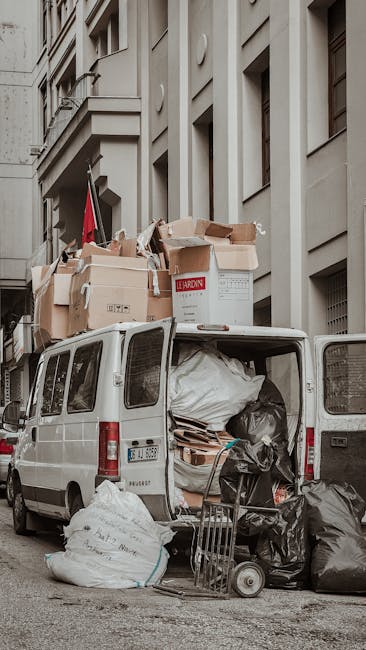 The image shows the open rear of a white van parked on a city street, loaded with various packing materials, cardboard boxes, and plastic-wrapped items for a home relocation. The van's interior is filled with large black rubbish bags, some wrapped furniture parts, and flattened cardboard sheets, indicating an ongoing packing and moving process. Several boxes are stacked on top of each other, secured with packing tape and placed inside the van, with some boxes marked for easy identification. A trolley is positioned nearby on the pavement, ready to assist with moving furniture or boxes. The backdrop features a tall, beige stone building with multiple windows and a flag hanging from a balcony. The setting suggests a professional removals service, such as Removal Van Kingston, involved in furniture transport and packing for a residential move, emphasizing organized logistics and careful handling of belongings during the loading process.