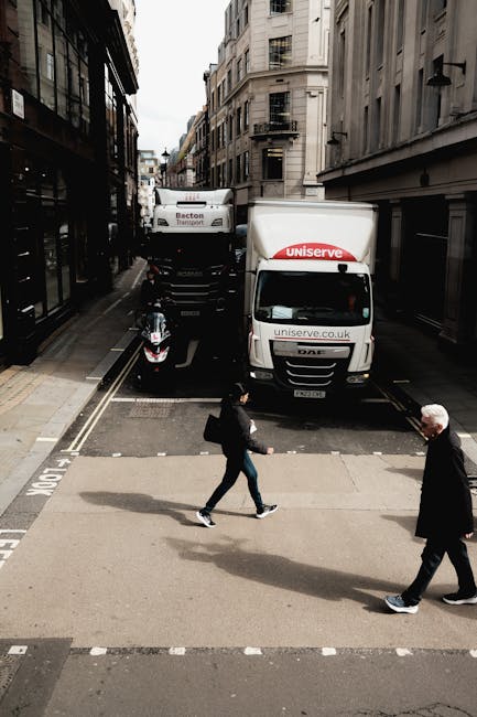 A narrow city street in Kingston, lined with multi-story stone and brick buildings, featuring a loading zone with two parked white removal vans branded with 'uniserve.co.uk' and 'Becton Transport.' The loading area contains a black motorcycle parked beside the vans, and a small, wheeled platform or trolley is situated near the vehicles. Two pedestrians are crossing the street at a zebra crossing; one is a woman dressed in dark clothing holding a black bag, and the other is an older man with white hair wearing a black jacket and dark trousers. The scene captures a typical urban home relocation environment with the vans positioned close to the building entrances, indicating a furniture transport or packing and moving process by Removal Van Kingston, part of professional removals services.