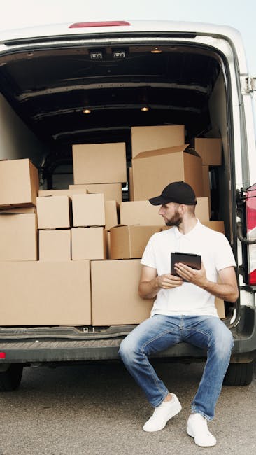 A young man with light skin, short light brown hair, and wearing a blue zip-up hoodie, navy trousers, and white sneakers, is seated on the edge of the open back of a white moving van. He is holding a