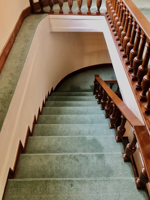 A downward view of a carpeted staircase inside a residential property, showing a series of green carpeted steps with wooden edges, leading to a lower floor. The staircase is flanked by a wooden handra