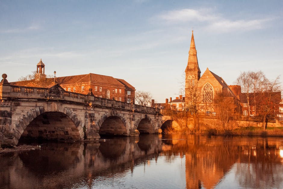 A wide view of a historic stone bridge with multiple arches spanning a calm river, reflecting the structure and surrounding buildings. In the background, there are several brick and stone buildings, including a prominent church with a tall, pointed spire, and smaller structures with pitched roofs. The scene is bathed in warm, golden light typical of late afternoon or early evening, highlighting the textures of the stone and brick surfaces. The riverbank has some leafless trees, indicating a season outside of summer. Near the river, a moving van from Removal Van Kingston is visible, with furniture and packed boxes inside, positioned near the property for home relocation activities. The van's open back doors suggest a loading or unloading process involving cardboard boxes, wrapped furniture, and moving equipment such as trolleys and straps, typical of professional removals during house moves in Kingston area. The overall scene captures a moment of furniture transport and packing during a local house removals service in Kingston.
