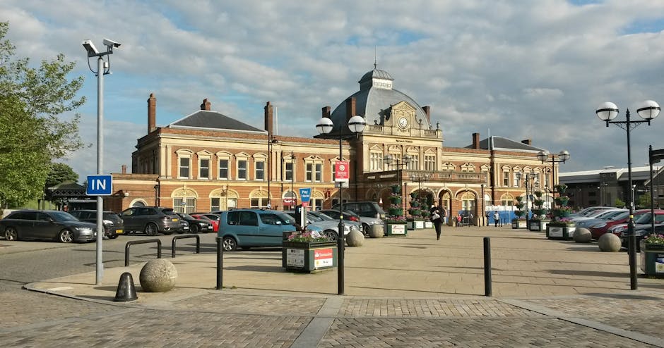 Photograph of Norbiton station's exterior in Kingston, showing a historic brick building with a central clock tower and decorative architectural details. The station is surrounded by a spacious paved area with parked cars, black metal bollards, and lampposts with spherical lights. A few pedestrians are visible walking near the building, and there are flower planters along the pavement. The scene is lit with natural daylight and a partly cloudy sky overhead. This setting captures the environment where home relocation and furniture transport services, such as those offered by Removal Van Kingston, often begin or conclude during house removals and packing and moving processes.