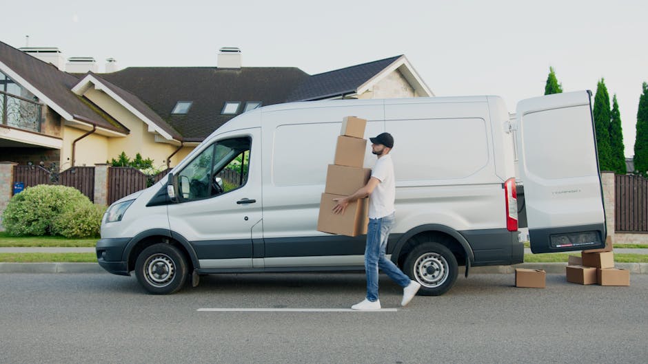 A man wearing casual clothing and a face mask is loading three cardboard boxes into the side door of a white commercial van parked on a residential street. The van, branded with neutral grey accents, is positioned next to a sidewalk and in front of a modern house with a dark roof, beige walls, and multiple attic windows. Additional boxes are placed on the pavement near the van’s open rear door. Inside the vehicle, visible through the open side door, are various packing materials such as plastic wraps and blankets, suggesting a home relocation or furniture transport process. The street is lined with well-maintained gardens and tall green trees, with clear weather and natural daylight illuminating the scene. The image reflects the loading and packing stage of a professional removals service by Removal Van Kingston, focusing on efficient logistical handling during a house move or apartment relocation.