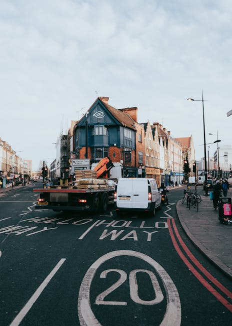 A flatbed truck operated by Removal Van Kingston is parked in the middle of a busy urban street in Kingston, loaded with wooden pallets, cardboard boxes, and wrapped furniture materials for a home relocation. The truck is positioned adjacent to a white van, likely assisting with the furniture transport and packing process for an upcoming house move. The roadway features visible traffic markings, including a 20 mph speed limit zone, with double red lines along the curb indicating restricted parking. Surrounding the scene are commercial buildings with multiple stories, some with decorative brickwork and bay windows. Pedestrians are walking on the pavement, and bicycles are parked nearby, suggesting a lively, accessible environment. Streetlights and signage are present, with light weather conditions and a partly cloudy sky overhead, reflecting typical city street conditions for local household removals and relocation services by Removal Van Kingston.