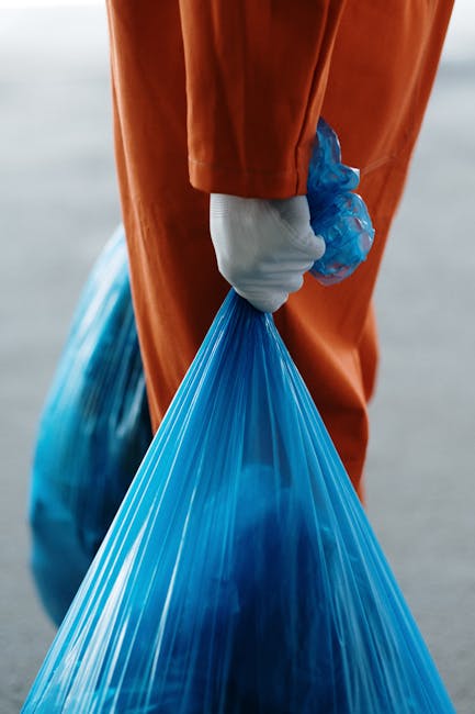A person wearing orange work overalls and white shoes is holding two large blue plastic bags filled with waste materials, standing on a grey concrete floor against a plain white wall. The individual i
